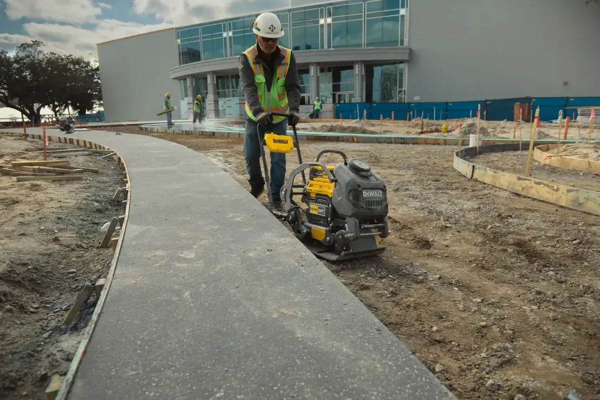 A worker running a plate compactor along a fresh concrete path beside a modern building — concrete business insurance