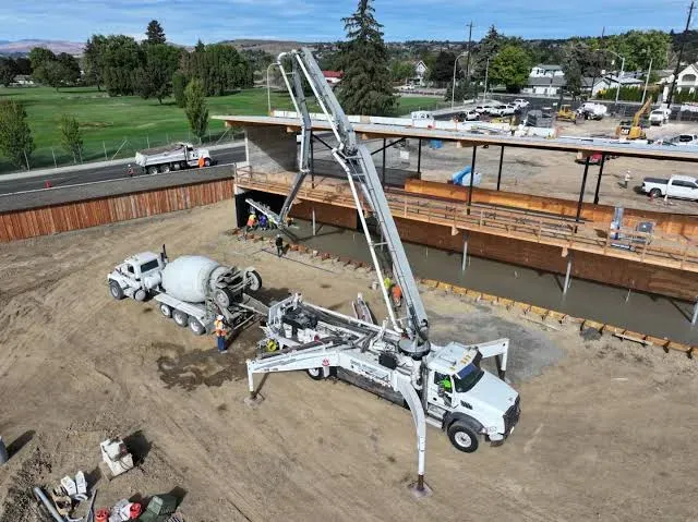 A boom pump set up on outriggers placing concrete beneath a bridge deck, fed by a mixer truck — concrete pumping insurance in Utah